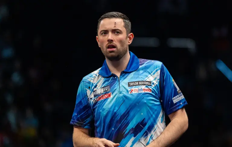 Luke Humphries bei der Swiss Darts Trophy 2026 in blauer Spielershirt auf der Bühne mit Dartpfeilen in der Hand.
