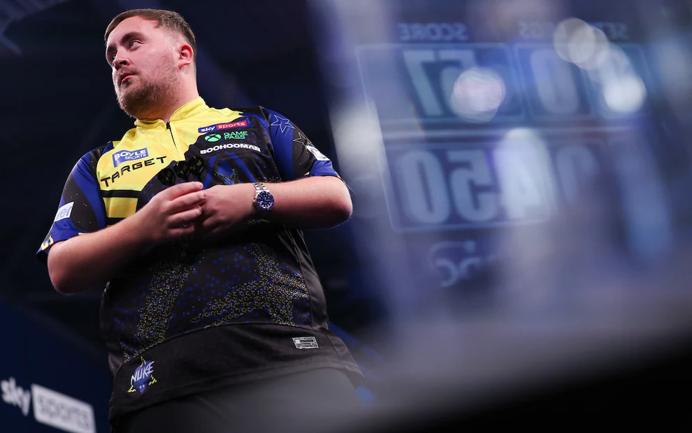 Luke Littler auf der Darts-Bühne während eines Turniers, mit Darts in der Hand und einer Anzeigetafel im Hintergrund. Kopieren